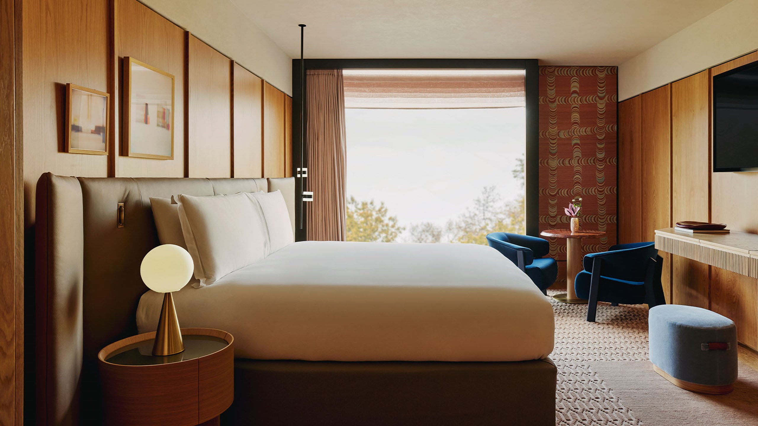 Bedroom of the Corner Park Suite, with a brown bed and white bedding, two blue armchairs, and views of Hyde Park out the floor to ceiling glass windows