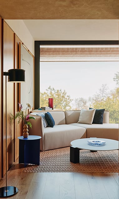 Cream sofa with dark blue cushions, placed in the sitting room of the Corner Park Suite, with large glass windows behind revealing leafy green trees