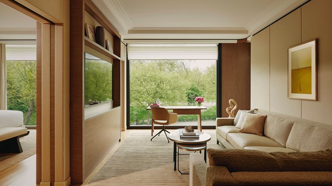 Contemporary living area with neutral-toned sofa, layered coffee tables, desk and chair by a picture window, and built-in TV with surrounding wood accents.