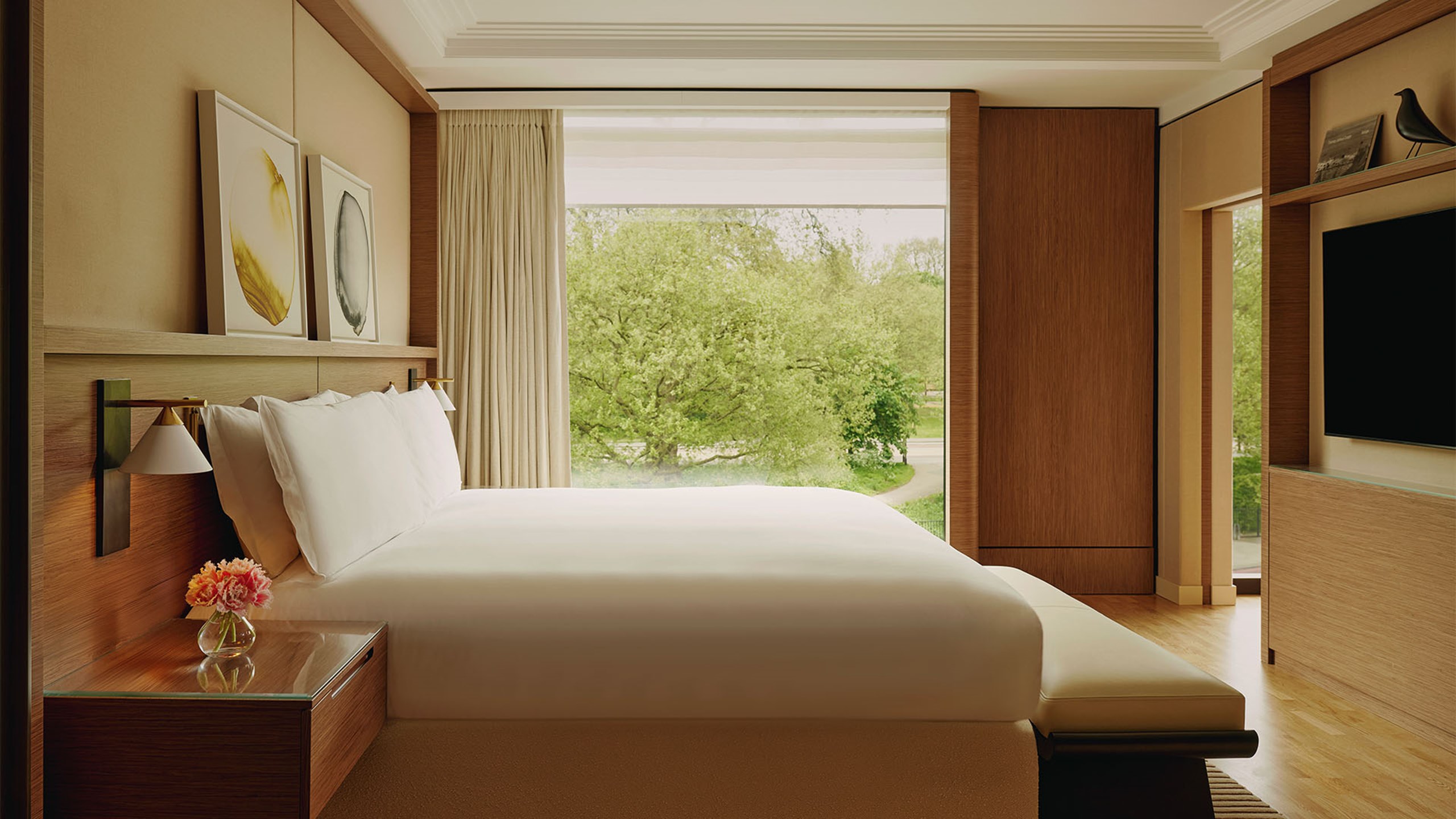 Bright bedroom with large bed, modern wall sconces, abstract artwork, and floor-to-ceiling windows offering serene views of green parkland and a winding path.