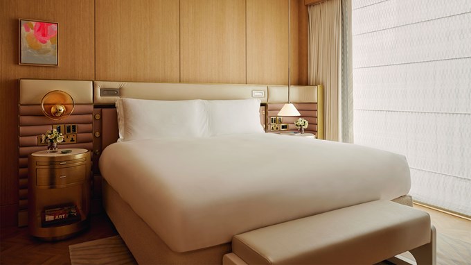 Courtyard Junior Suite bedroom. A window is to the back right next to a round glass table and two chairs in the right foreground. A double bed sits in the foreground with a bench at the end of the bed. Two marble and wooden bedside tables are located next to the bed.