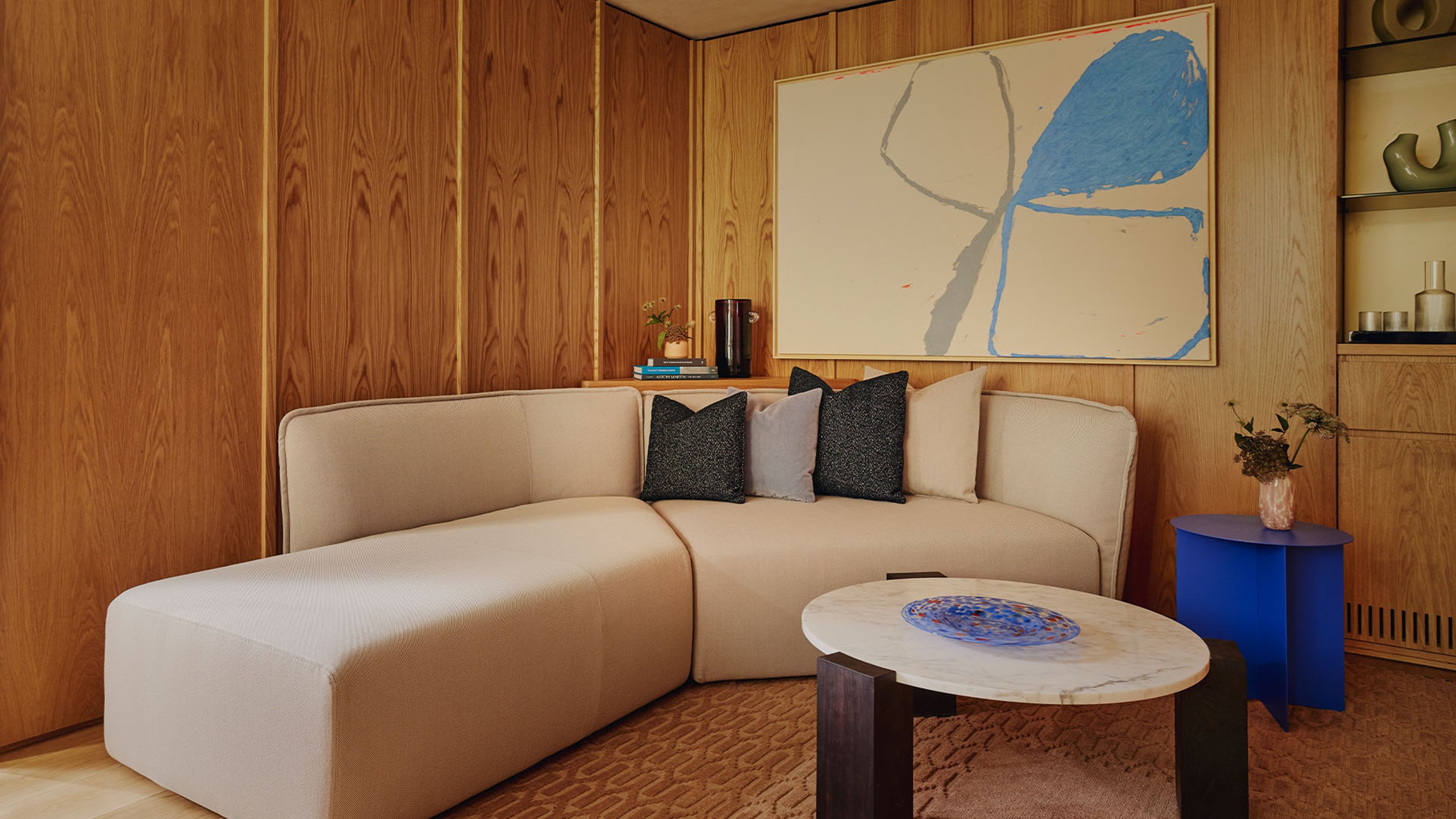 Courtyard Junior Suite sitting area. A long beige couch sits in the middleground and a circular wood and marble table sits in the foreground. A small round blue table sits behind it. A large painting and picture frame is hung on the wall behind the couch.