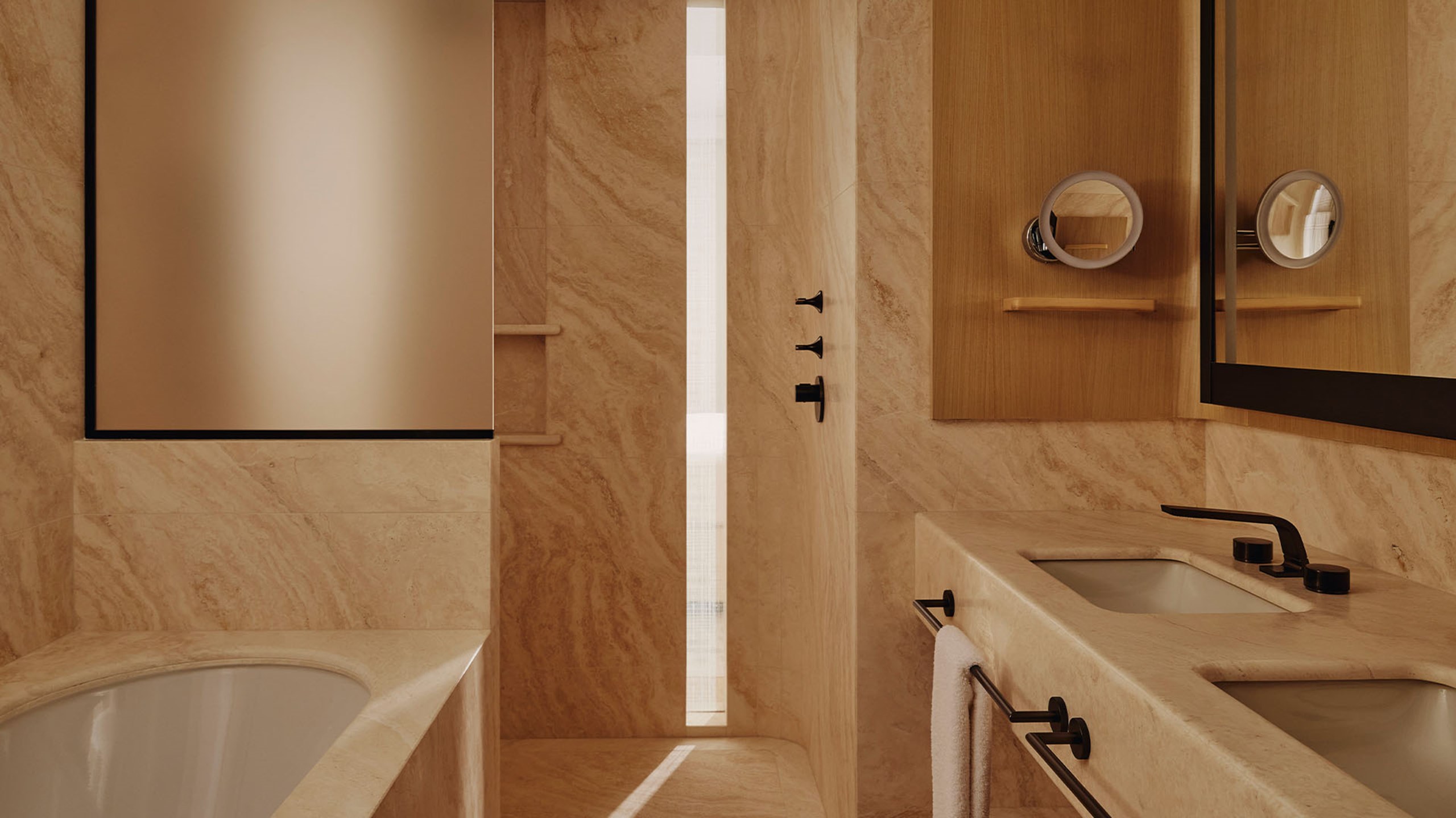 Courtyard Junior Suite marble bathroom. A large mirror is located on the wall to the right on top of two bathroom sinks. A towel rail sits below each sink. The entrance to the shower is located in the background. A bath is placed on the left.