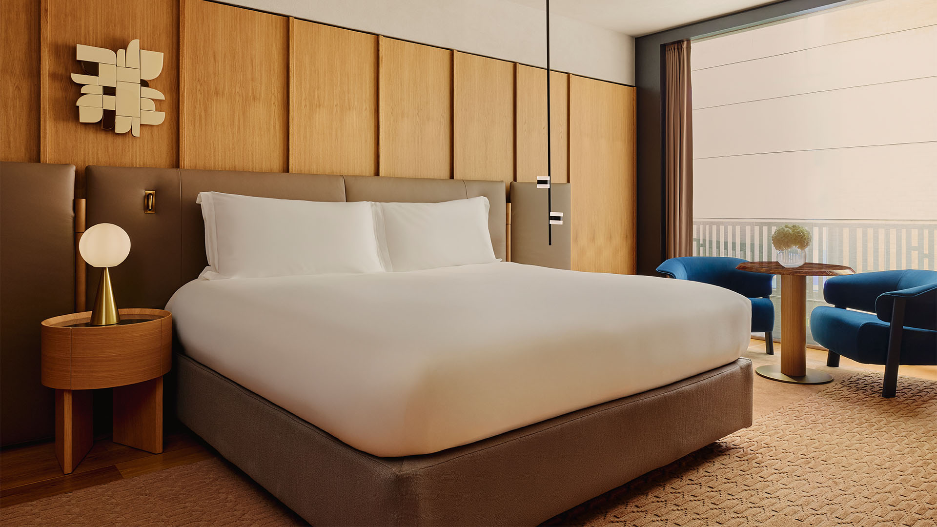Warm bedroom with timber panels, large bed, round bedside lamp, and two blue armchairs by a window table.