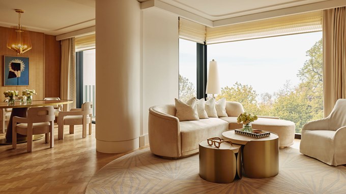 Spacious living area with bar and sitting area, with light pink, beige and gold decor. In the foreground is a coffee table with stylish coffee table books and gold statement pieces on top, and in the background is glass windows looking out to the park and trees
