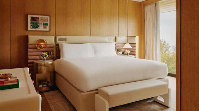 Wooden panels on walls of bedroom of Deluxe Balcony Park Suite. The light coloured bed has white bedding, gold lamps on bedside tables and glass windows revealing the trees outside.
