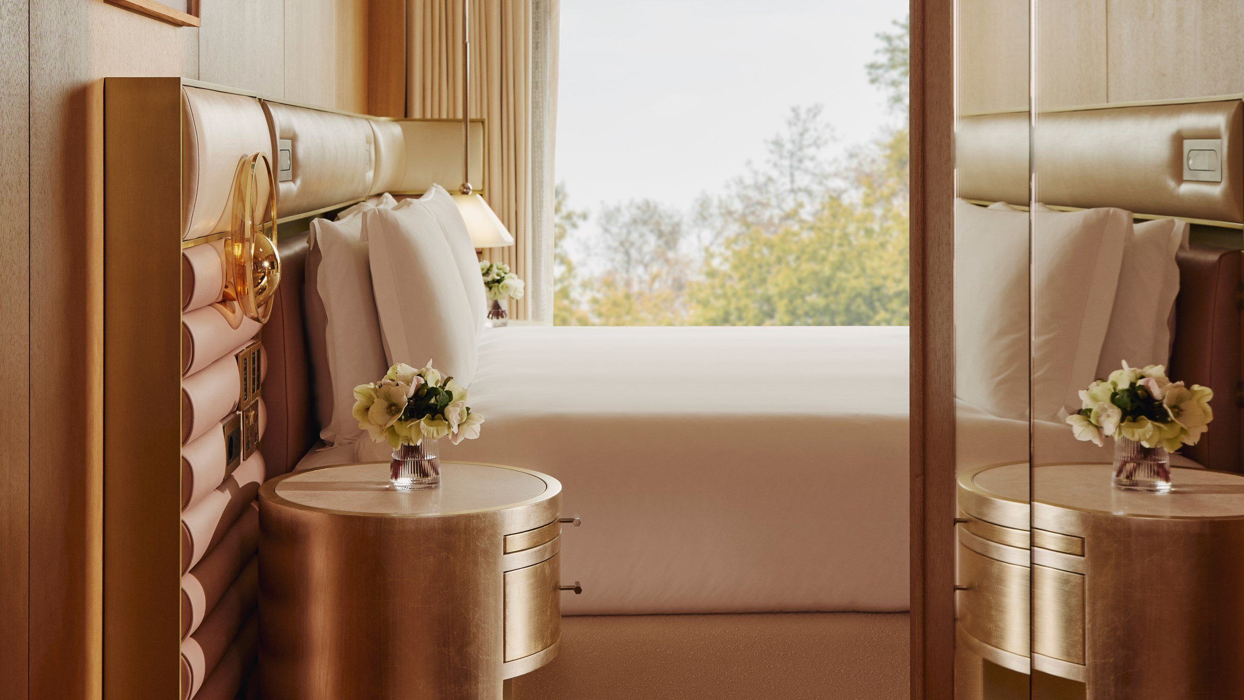 The Emory Deluxe Balcony Park Suite Bedroom - A window sits in the background and a double bed sits in front of it. A round gold bedside table with white flowers in a vase sits next to it. A mirror sits to the right.