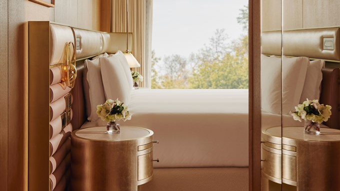 The Emory Deluxe Balcony Park Suite Bedroom - A window sits in the background and a double bed sits in front of it. A round gold bedside table with white flowers in a vase sits next to it. A mirror sits to the right.