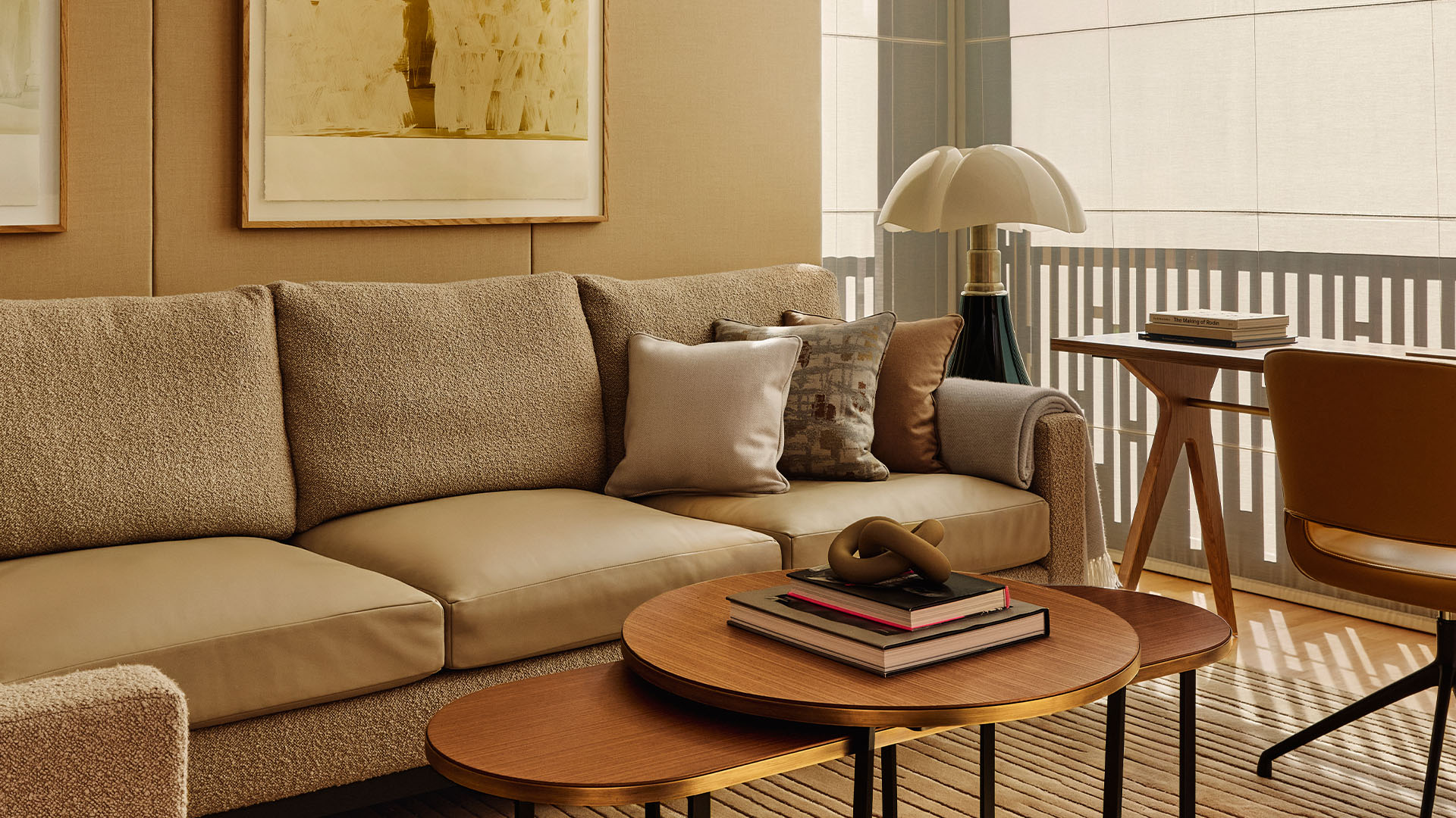 Neutral-toned lounge with textured sofa, layered coffee tables, desk area, and soft light through blinds