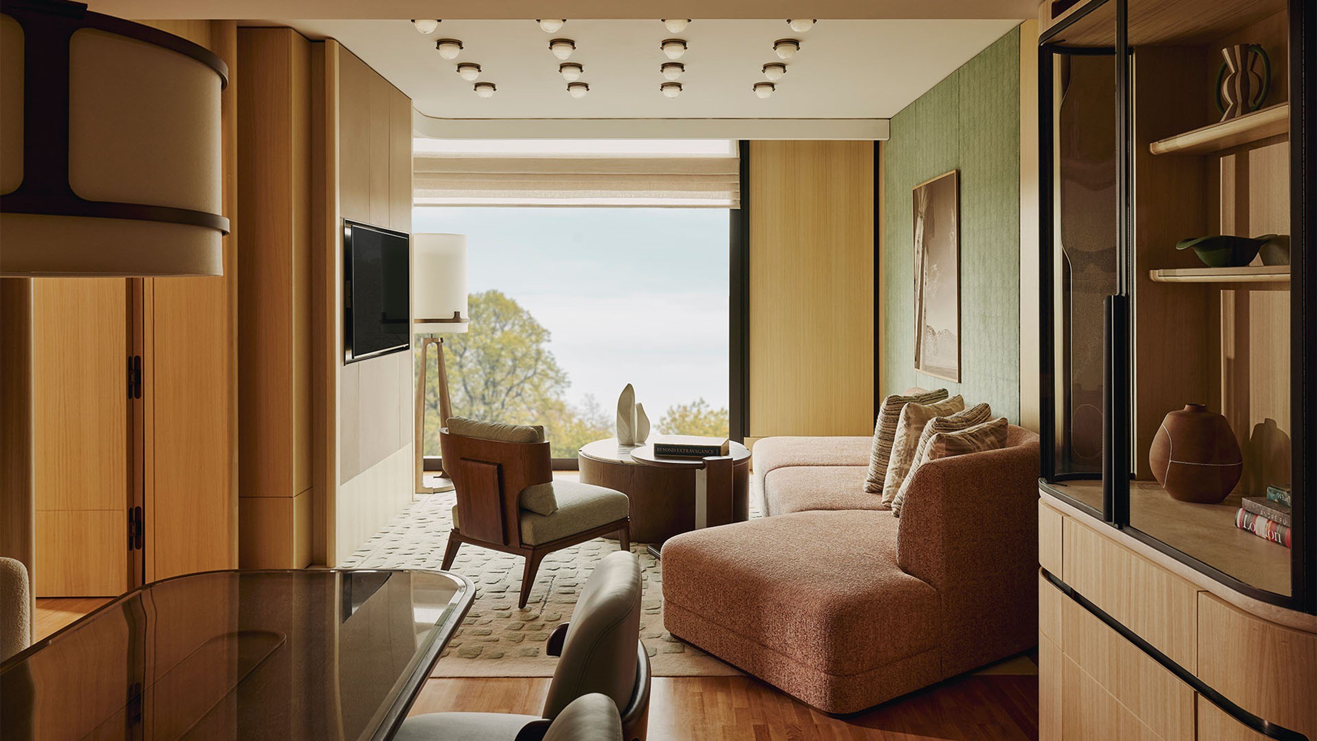 Contemporary living area with neutral-toned sofa, layered coffee tables, desk and chair by a picture window, and built-in TV with surrounding wood accents.