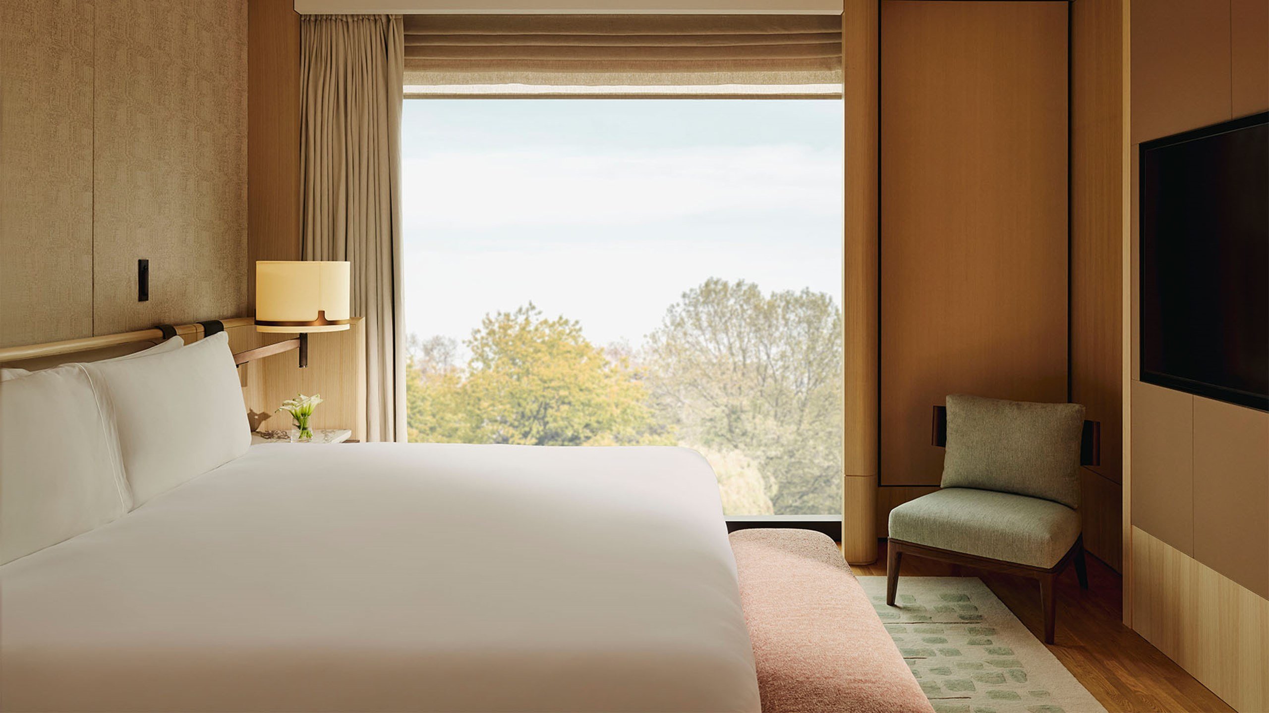 Bright bedroom with large bed, modern wall sconces, abstract artwork, and floor-to-ceiling windows offering serene views of green parkland and a winding path.