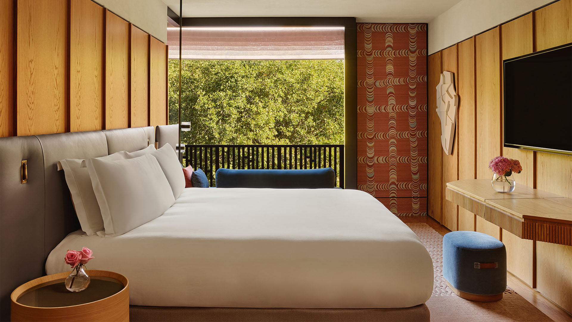 Bright bedroom with a large bed, wood panelled walls, blue bench, and balcony view of green trees