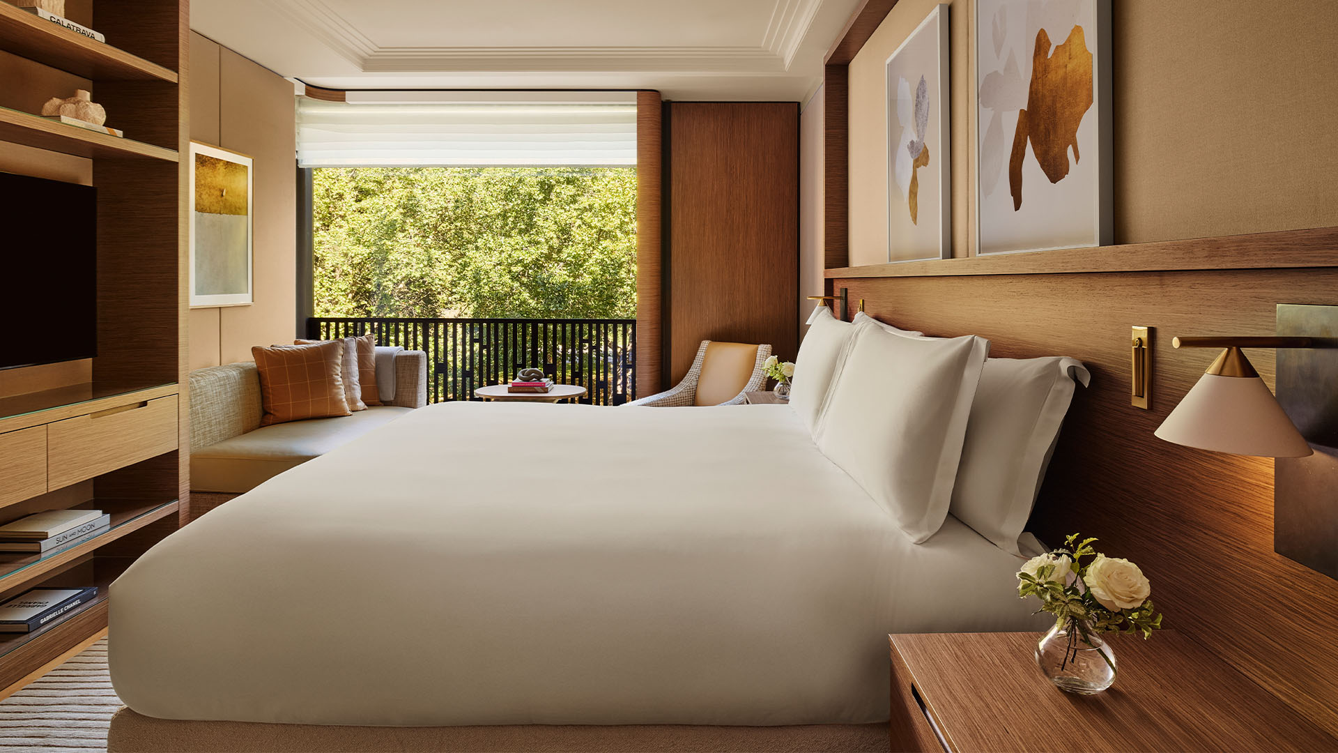 Warm-toned bedroom with large bed, built-in sofa, framed art, and balcony overlooking trees.