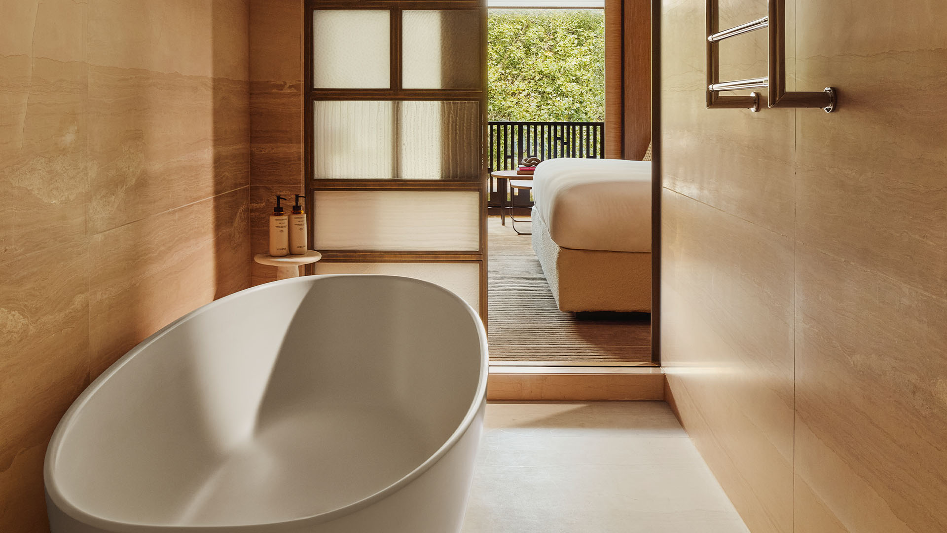 Stone-tiled bathroom with freestanding tub, glass screen, and view into the bedroom and balcony.