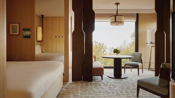 Park Junior Suite bedroom and sitting area. A bed sits to the left and a mirror behind it. A chair sits to the right. A circular wooden table sits in the background and a couch and chair sit around it. A large window sits behind the table.