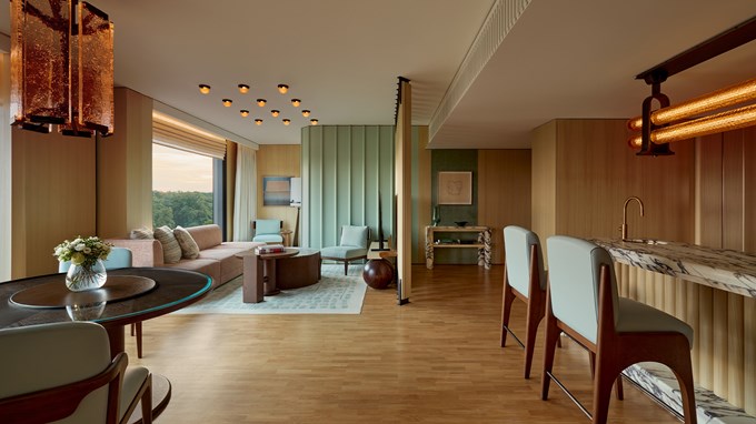 A spacious, elegantly furnished suite with soft wooden tones, a marble-topped bar, and floor-to-ceiling windows offering a tranquil view of lush greenery.