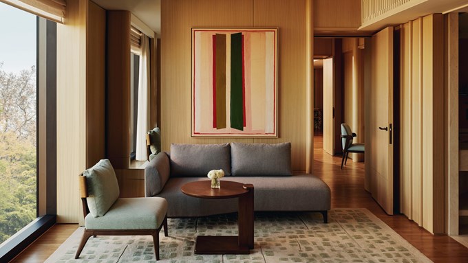 The Emory Park Suite Lounge Area - A window sits to the left. A green and wooden chair sits in front of it. A round wooden table with flowers in a vase sits in the middle ground. A grey couch sits behind it. A brown, green and pink painting is hung on the wall behind the couch.