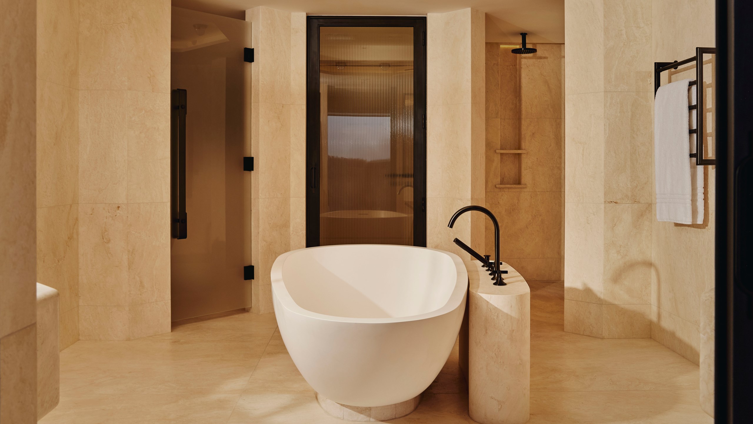 The Emory Park Suite Bathroom - A white bath sits in the foreground. A wooden and glass door is behind it. A marble shower sits in the right background. A white towel is hung to the left.