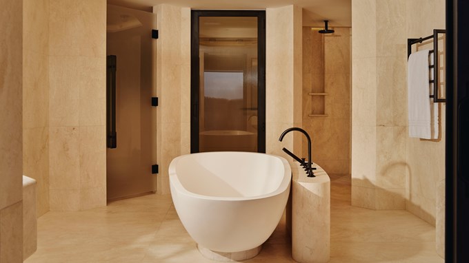 The Emory Park Suite Bathroom - A white bath sits in the foreground. A wooden and glass door is behind it. A marble shower sits in the right background. A white towel is hung to the left.