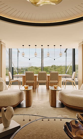 Light-filled living and dining space with curved cream seating, wood tables, and floor-to-ceiling windows overlooking trees.