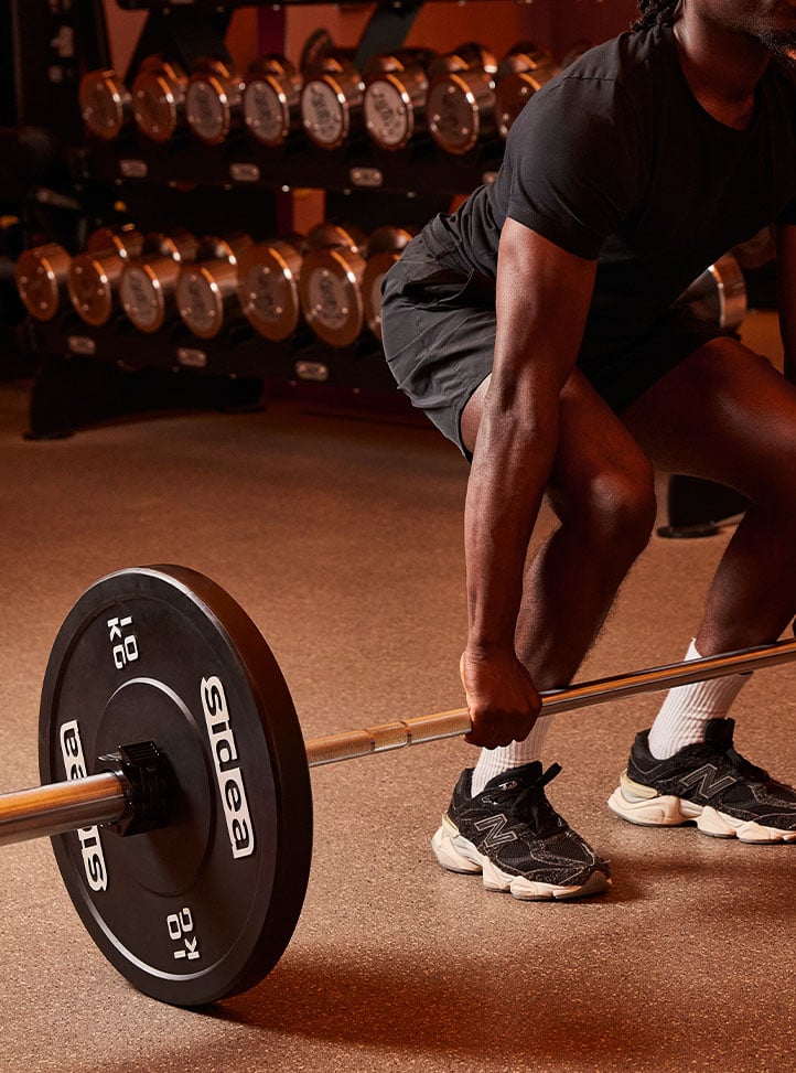 A man wearing black athletic clothing and trainers prepares to lift a barbell in a gym, with a rack of chrome dumbbells in the background