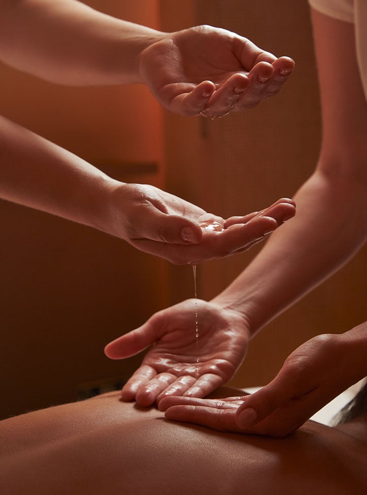 A spa guest in a black one-shoulder top lies on a treatment bed with eyes closed, while a therapist places their hands gently on her forehead in a softly lit room at Surrenne Spa, The Emory.